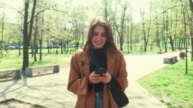 Genç mutlu bir kadının bahar zamanı parkta yürüdüğü ve akıllı telefondan yazı yazdığı görüntüler..