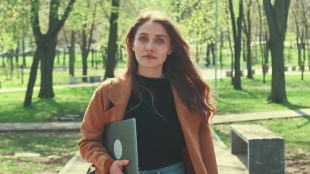 ノートパソコンを持って街の公園を歩く若い女性の動きビデオは 仕事に行きます — ストック動画