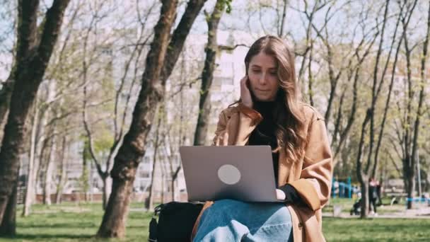 若いです魅力的な女性のビデオ座っている屋外で公園と作業ノートパソコン — ストック動画