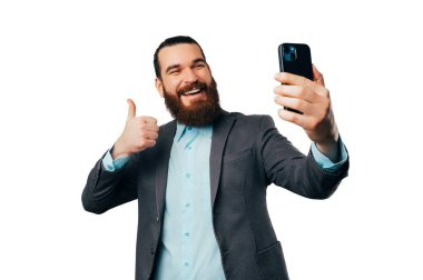 Yakışıklı sakallı adam beyaz arka planda baş parmağını gösterirken telefonuyla selfie çekiyor..