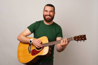 Neşeli genç sakallı adam akustik bir gitarla tutuyor ve çalıyor. Stüdyo resmi.
