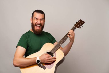 Kendinden geçmiş genç sakallı adam gri arka planda akustik bir gitarla rock çalıyor..