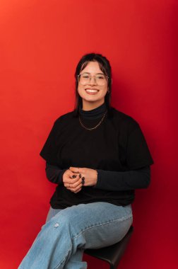 Kırmızı arka planda bir sandalyede oturan gülümseyen genç bir kadının dikey stüdyo çekimi..