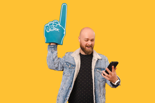 Studio portrait of a cheerful bearded bald man wearing foam finger glove and watching soccer on the phone.