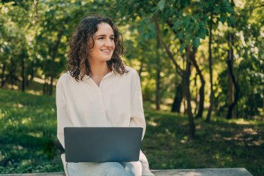 Neşeli kıvırcık kadın dizlerinin üstünde bir dizüstü bilgisayar tutuyor ve parkta oturuyor..