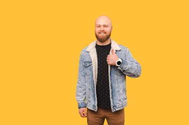 Kot ceketli, sırıtan kel sakallı adam sarı arka planda baş parmağını gösteriyor..