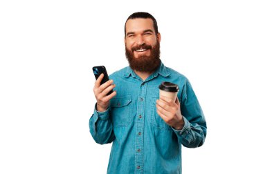 Neşeli genç sakallı adam elinde bardak ve telefonu için bir kağıt tutuyor. Stüdyo resmi.