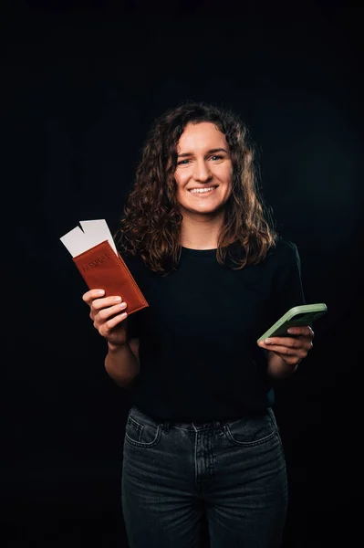 Stock image Vertical shot of a smiling woman holding her passport and smart phone over black background.