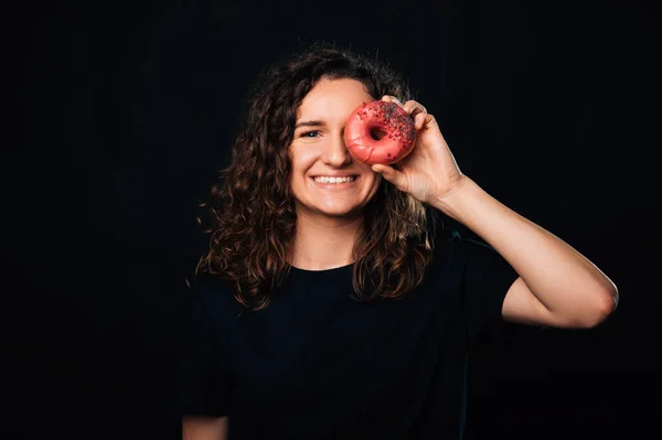 Wide smiling curly woman covers one eye with a delicious big pink donut over black background.