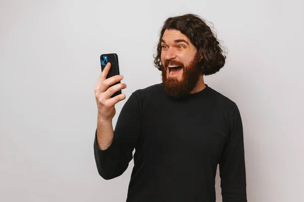 stock image Young bearded man with long hair is looking shocked at the phone he is holding.