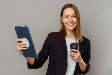 Ceket giyen neşeli kadın elinde tablet ve gri arka planda bir fincan kahve tutuyor..