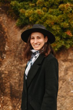 Açık havada siyah şapka ve ceket giyen gülümseyen genç kadının dikey portresini kapat..