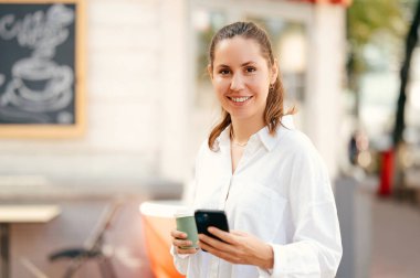 Güzel, genç bir kadın elinde telefonla kameraya gülümserken kahvesinin tadını çıkarıyor..