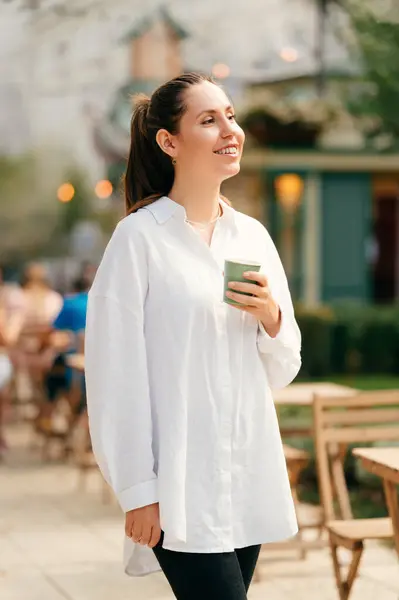 Vertical shot of a smiling confident woman enjoying her morning coffee on city streets.