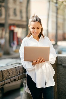 Beyazlı neşeli genç kadın balkonda dizüstü bilgisayarından e-postalarını kontrol ediyor..