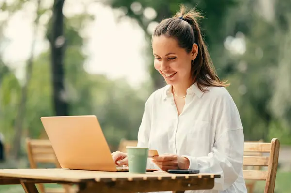 Smiling woman is using web banking or buying online with her card and laptop in a cafe in park.