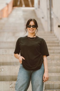 Merdivenlerde dururken güneş gözlüğü takan gülümseyen genç bir kadının dikey portresi.