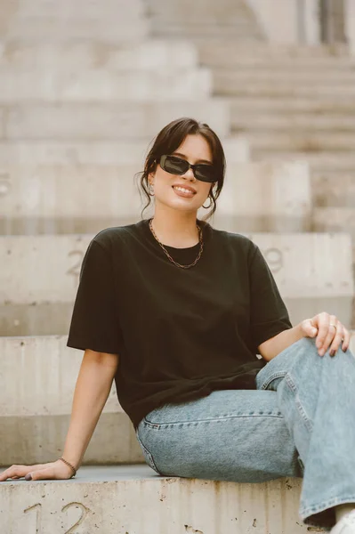 Cute young woman wearing black t shirt, jeans and sunglasses sits on concrete stairs.