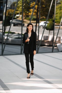 Dizüstü bilgisayarı olan ve yeni iş binasının yanında yürüyen genç bir kadın resmi.