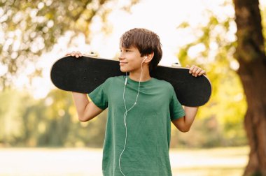 Kulaklıkla müzik dinleyen ve parkta kaykay tutan bir çocuğun fotoğrafı..