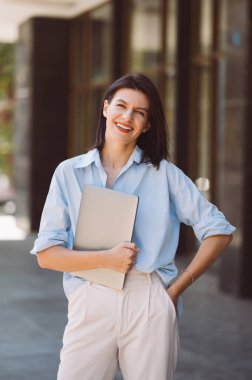 Gülümseyen genç iş kadını dizüstü bilgisayarını tutuyor ve kameraya bakıyor...