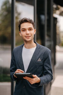 Genç kendine güvenen bir adam elinde dijital tabletle iş cam binasının yanında yürüyor..