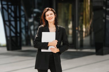 Şehrin dışında, yeni binanın yakınında elinde dizüstü bilgisayarla gezen neşeli genç bir iş kadını fotoğrafı..