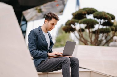 Konsantre genç adam dışarıda bir iş binasının yanında otururken dizüstü bilgisayarında daktilo yazıyor..
