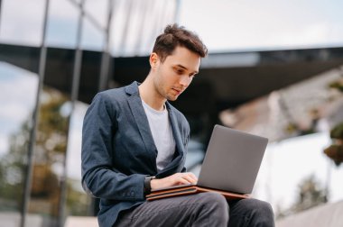 Yoğunlaşmış genç adam dışarıda cam bir binanın yanında otururken bilgisayarında daktilo yazıyor..