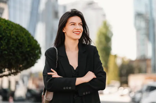 Nice young business woman wearing black jacket is walking outdoors with arms crossed.