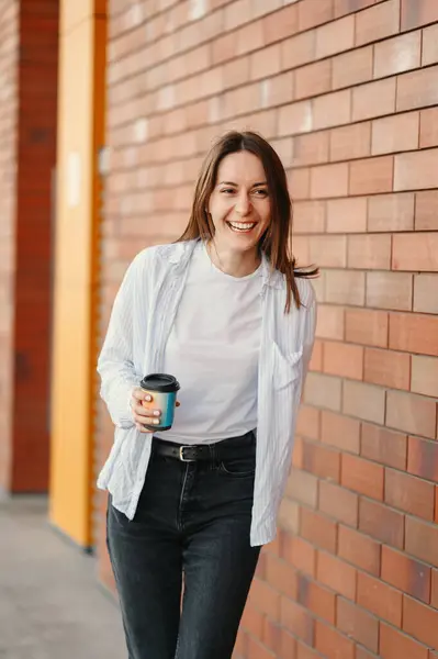 Vertical shot of a laughing young woman holding take away coffee while walking by a brick wall.