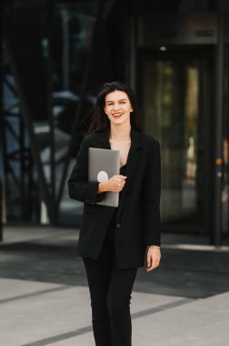 Kendine güvenen bir kadının siyah takım elbise giyip dışarıda dizüstü bilgisayar tutarken dikey görüntüsü..