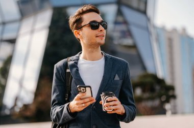 Ceketli, güneş gözlüklü, elinde kahve ve telefonu olan iyimser bir genç..