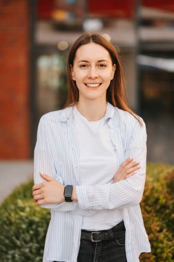 Beyaz elbiseli, kolları açık bir binanın yanında duran gülümseyen bir kadının dikey çekimi..