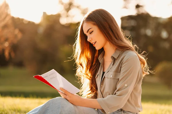 Neşeli genç kız öğrenciler parkta oturuyor ve güneşli bir günde bir kitaptan öğreniyorlar..