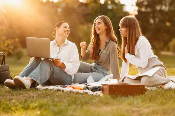 Three girls friends study at a picnic in park while watching an online course on laptop.