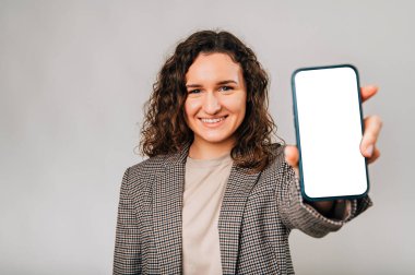 Ceketli, kendine güvenen genç bir kadın boş bir telefon ekranı gösterirken sırıtıyor..