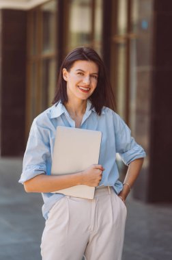 Güneşli bir günde tişört giyen ve dizüstü bilgisayar tutan gülümseyen bir kadının dikey portresi..