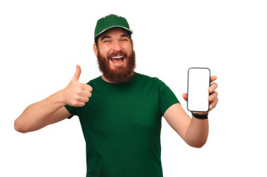 Yeşil üniformalı neşeli sakallı adam boş bir telefon ekranı ve başparmağını kaldırmış..