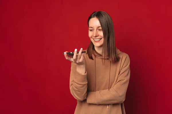 Cheerful young woman is talking on phone speaker ar making commands on it. Studio shot over red background.