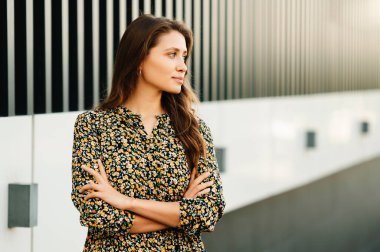 Açık havada, kollarını kavuşturup fotokopi alanına bakan genç bir kadının fotoğrafı..