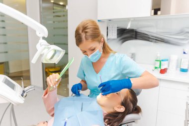 Genç bayan yıllık bir ziyaret ve konsültasyon için dişçide oturuyor. Doktor çalışırken o da aynaya bakıyor..