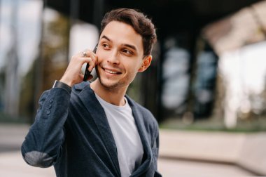 Ceket giyen yakışıklı genç bir adam telefon ederken yana bakıyor. Açık hava çekimi.