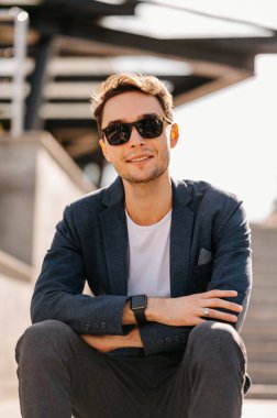 Ceket ve güneş gözlüğü takan kendine güvenen genç adam dışarıda kollarını kavuşturmuş oturuyor..