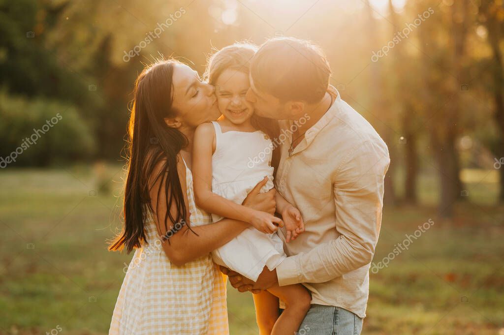 Happy daughter held up and kissed by her mother and father at sunset in park in summer.
