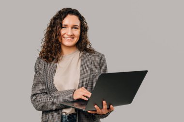 Ceket giyen kendinden geçmiş genç bir kadın elinde bir dizüstü bilgisayar tutuyor ve üzerinde çalışıyor. Stüdyo çekimi gri arkaplan üzerinde.
