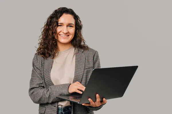 Ceket giyen kendinden geçmiş genç bir kadın elinde bir dizüstü bilgisayar tutuyor ve üzerinde çalışıyor. Stüdyo çekimi gri arkaplan üzerinde.