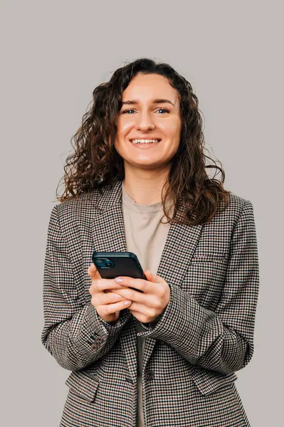 Geniş gülümseyen, ceketli ve gri arka planda telefonu tutan bir kadının dikey görüntüsü..