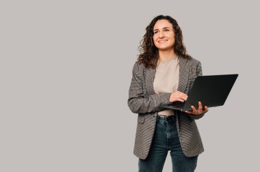 Ceket giyen kendinden geçmiş genç bir kadın hayal kurarken dizüstü bilgisayar tutuyor. Stüdyo çekimi gri arkaplan üzerinde.