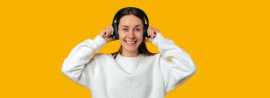 Heyecanlı genç bir kadının sarı arka planda kulaklık taktığı bir fotoğraf..